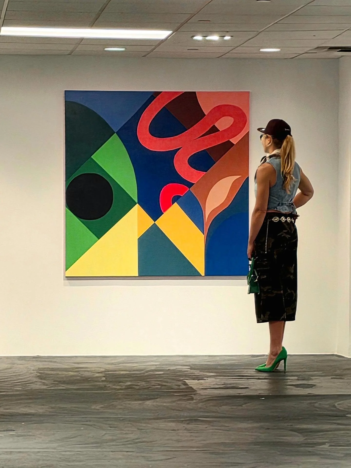 A woman with a ponytail, wearing sunglasses, a sleeveless denim vest, a black floral skirt, and green high heels, is standing in front of an abstract colorful painting in an art gallery.