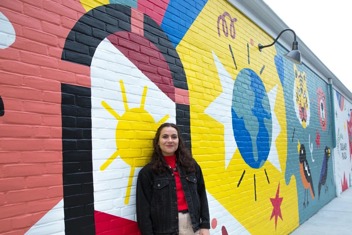 A woman standing in front of a colorful mural painted on a brick wall. The mural features various abstract shapes, a sun, and a globe.