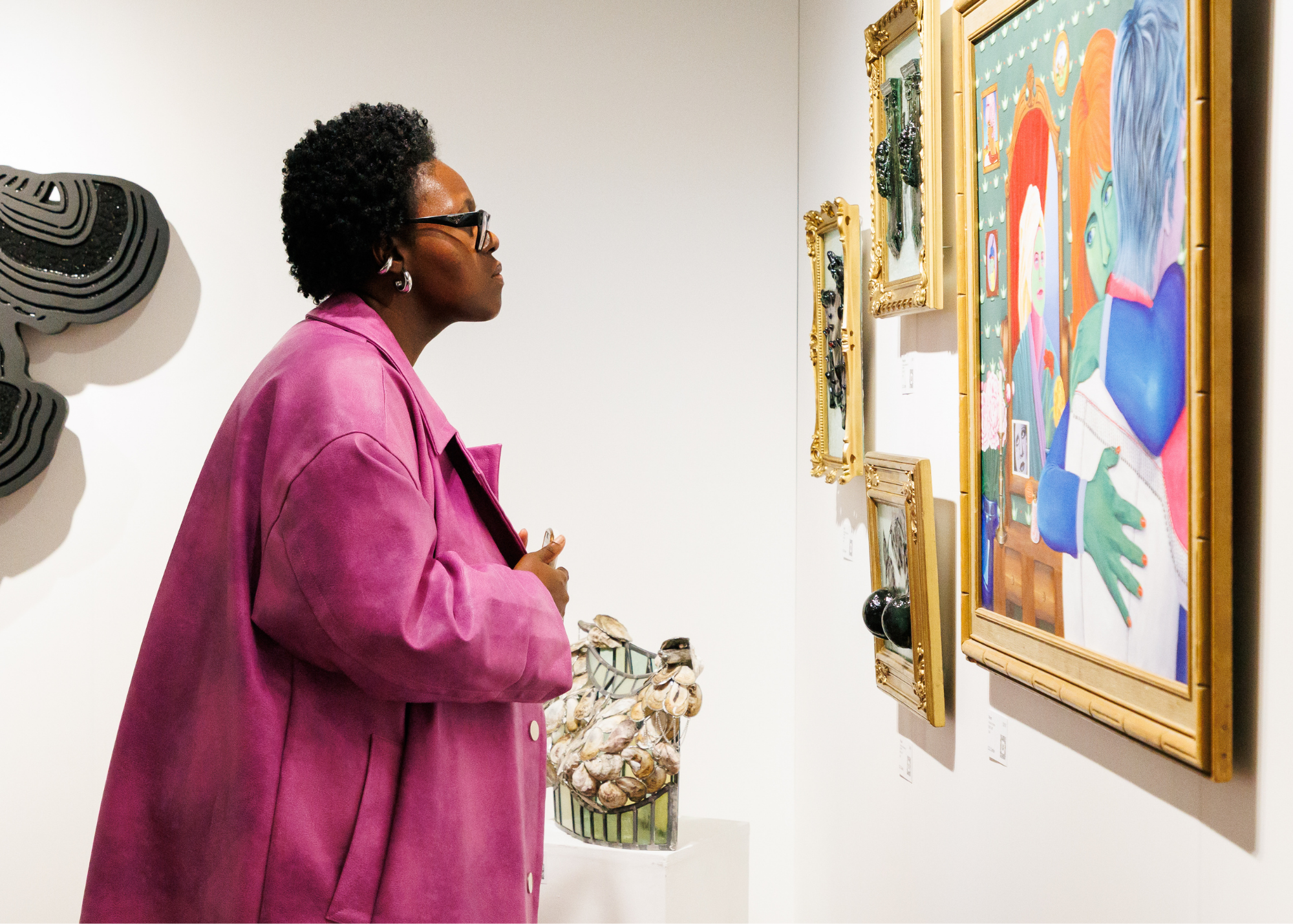 A woman with short, curly black hair and glasses wearing a pink jacket is observing colorful framed artwork in an art gallery.