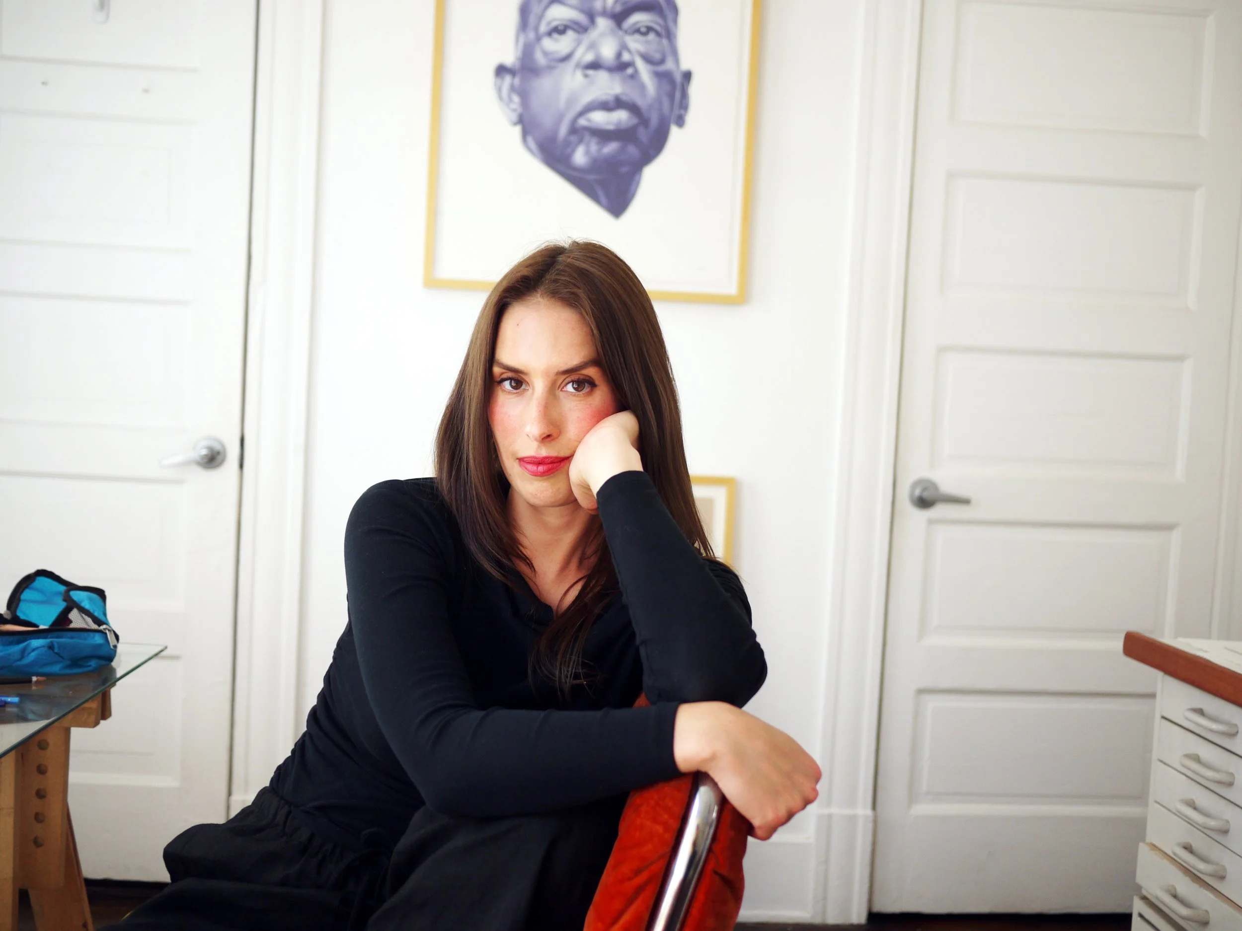 A woman with long brown hair and fair skin sitting at a table in a room with white walls and closed white doors. She is resting her head on her hand, looking at the camera. In the background, there is a large framed portrait of a man's face on the wall.