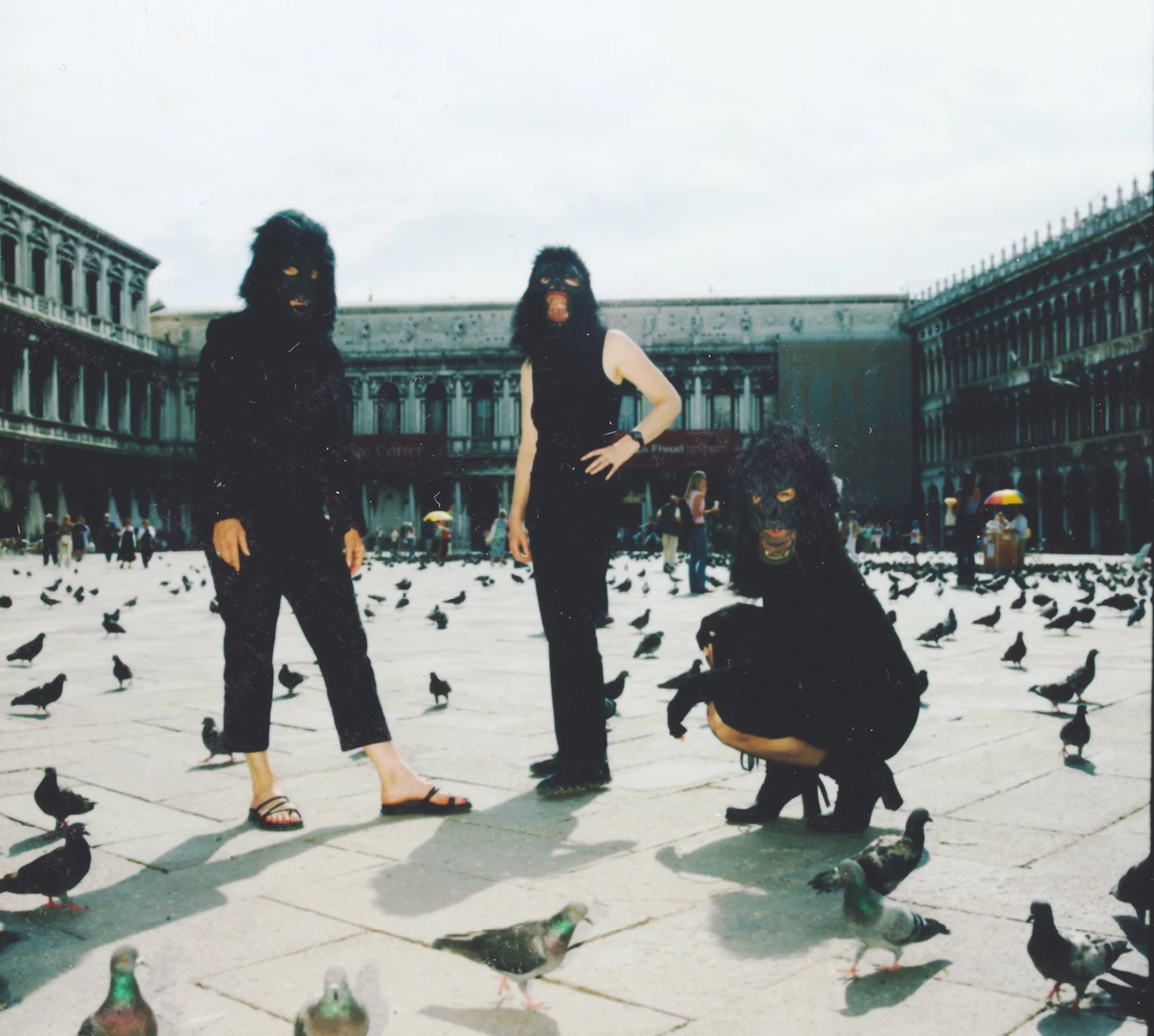 Three people with masks and black clothing in a pigeon-filled square, with historic architecture in the background and some onlookers.