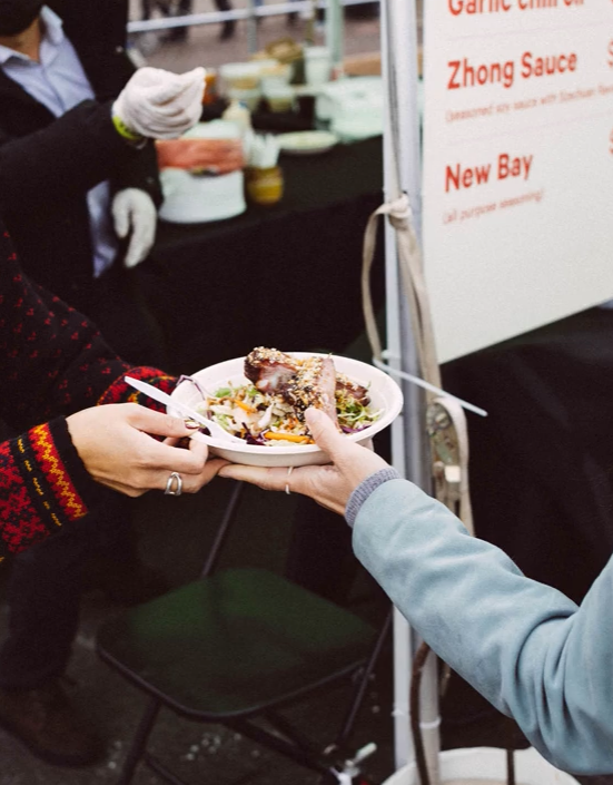 Someone serving a plate of food at a food stall, with a person receiving the plate aboard, at a market or fair.