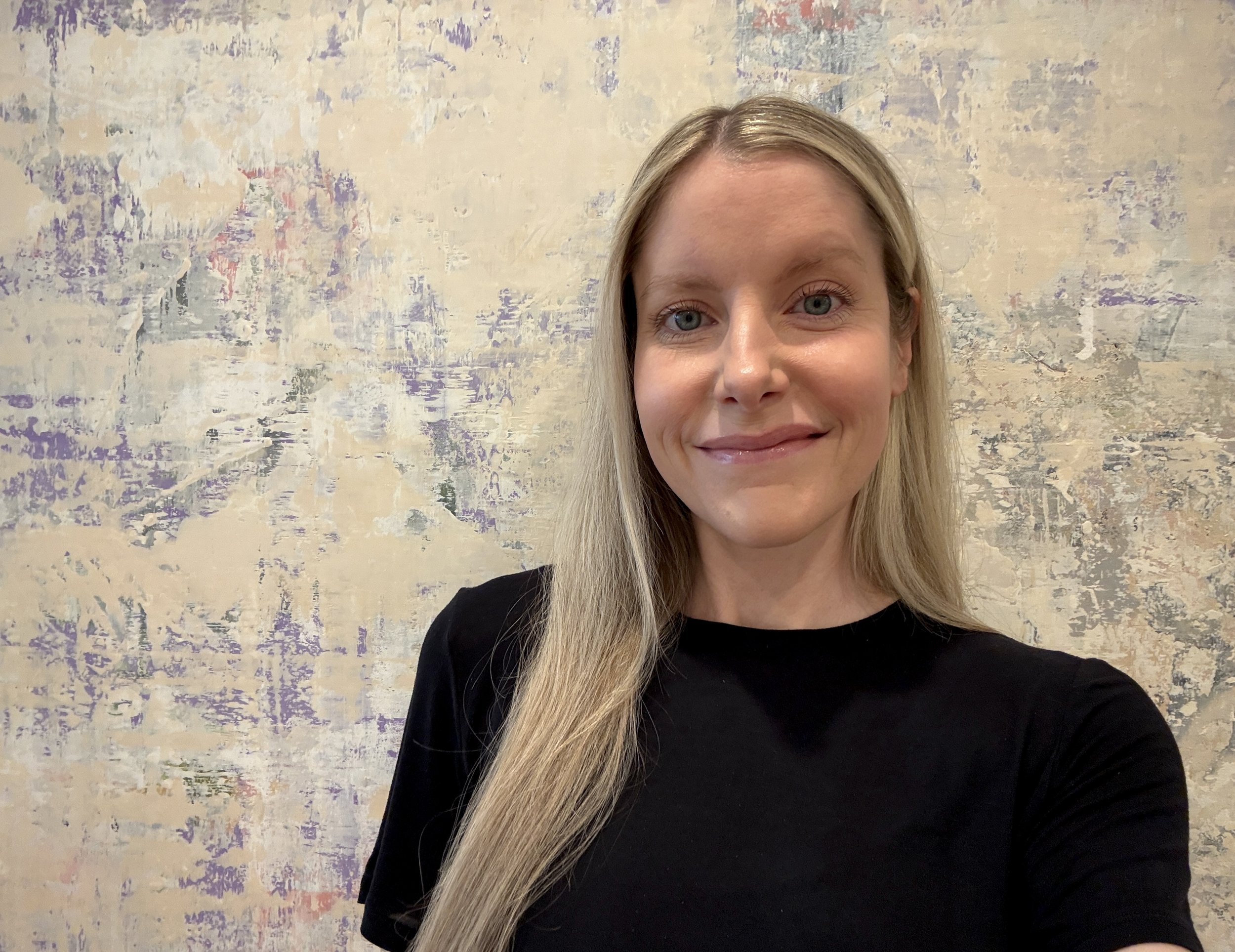 A woman with long blonde hair and blue eyes taking a selfie in front of an abstract textured wall.