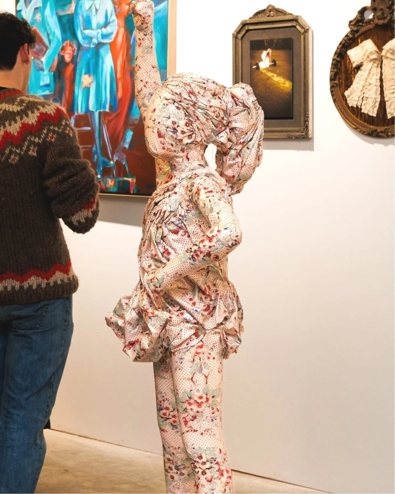 An art gallery shows a person in floral-patterned clothing with a large hood and baggy pants standing in front of artwork, while another person wearing a patterned sweater observes nearby.