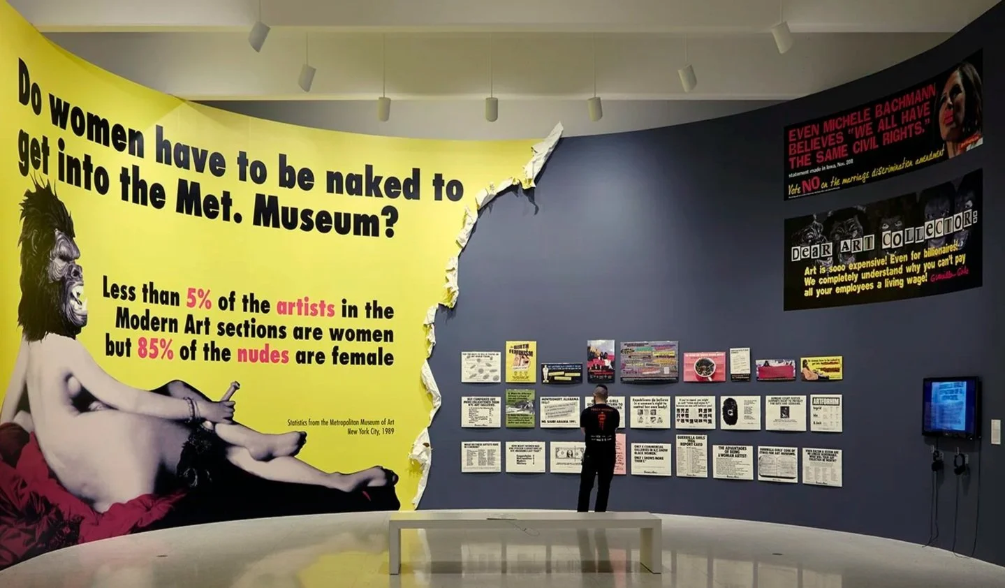 Exhibition space at the Metropolitan Museum of Art with a large yellow wall display questioning if women have to get naked to get into the museum. It states less than 5% of artists are women in modern art, but 85% of nudes are female, citing stats from 1989. The wall includes images and texts related to women artists and art history.