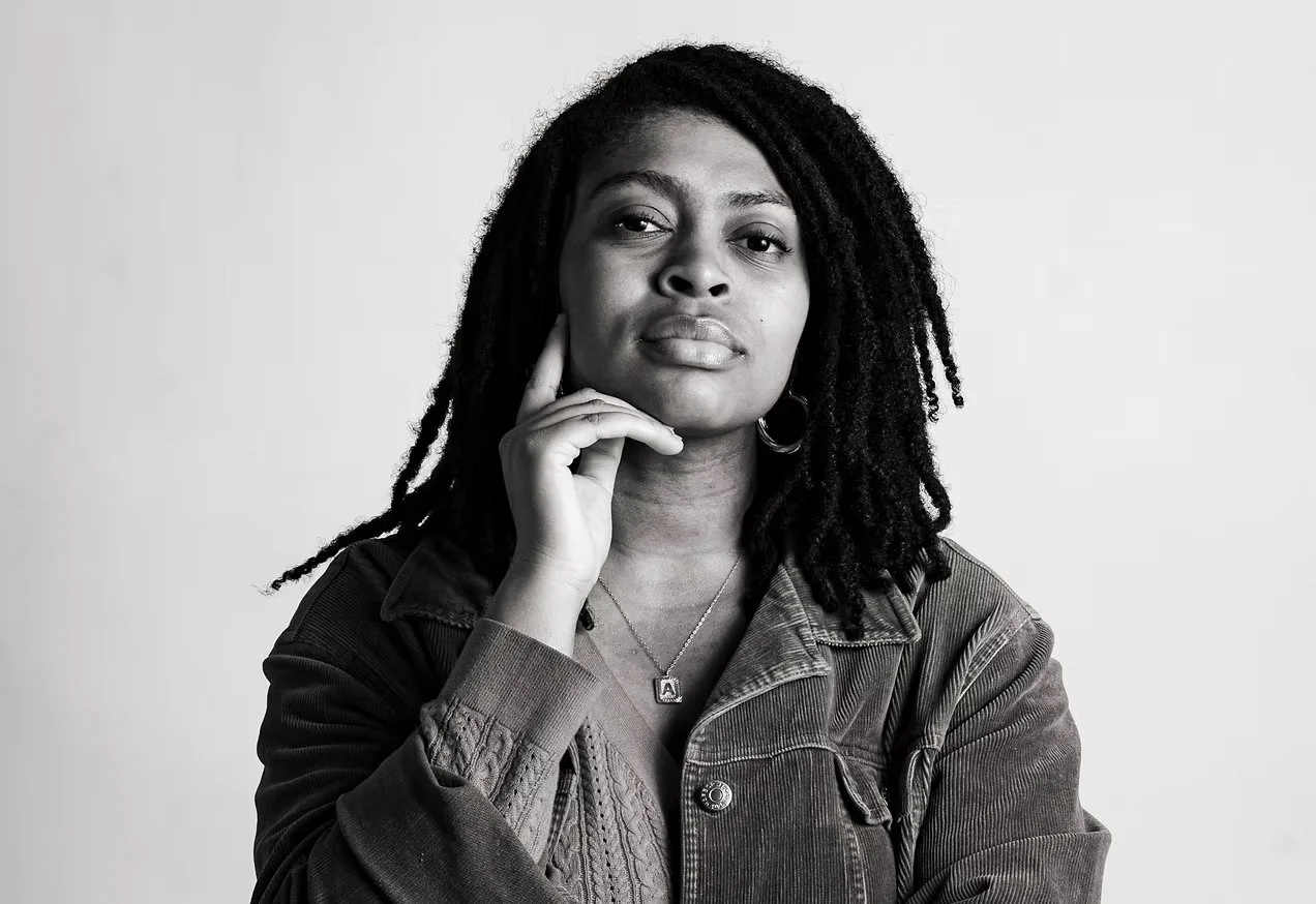 Black and white portrait of a woman with dreadlocks, wearing a denim jacket and a necklace with the letter 'A', posing with her chin resting on her hand.