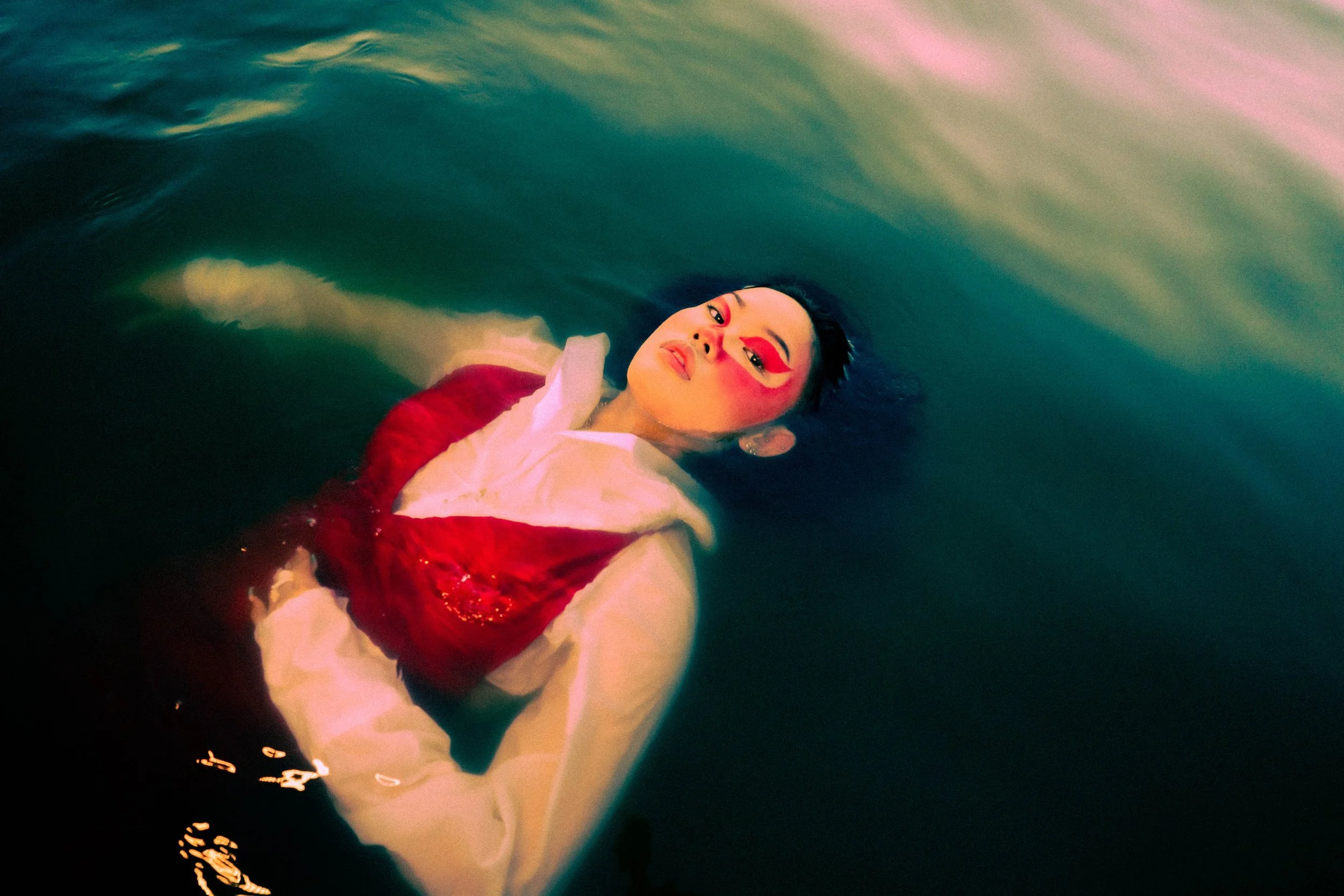 A woman with makeup lying in a body of water, wearing a red and white outfit, with colorful lighting creating a vibrant atmosphere.