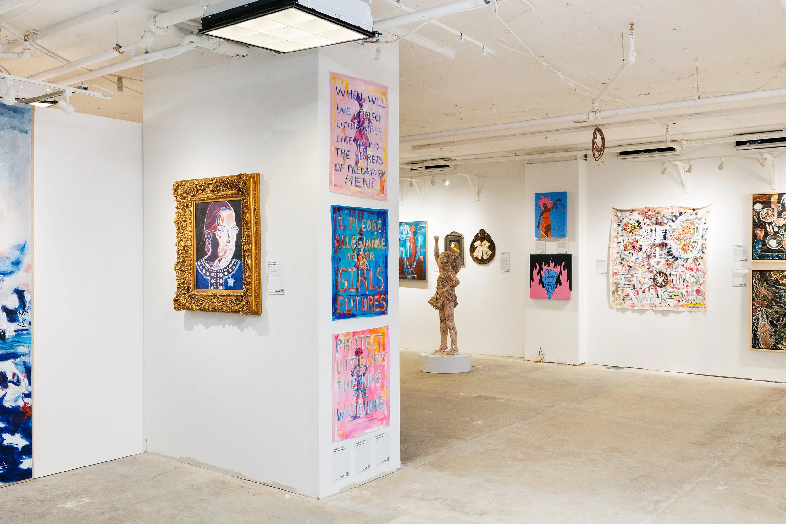 Art gallery displaying various contemporary paintings and mixed-media artworks, including colorful portraits, text-based art, and abstract pieces, in a white-walled space with polished concrete floors.
