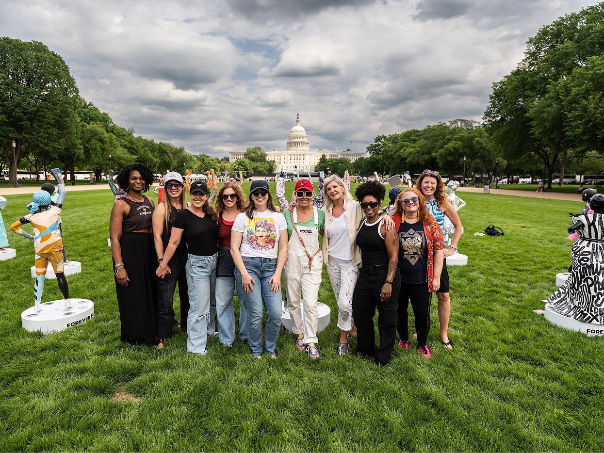 Group of women standing on grass field with sculptures in the background, U.S. Capitol building and cloudy sky visible.