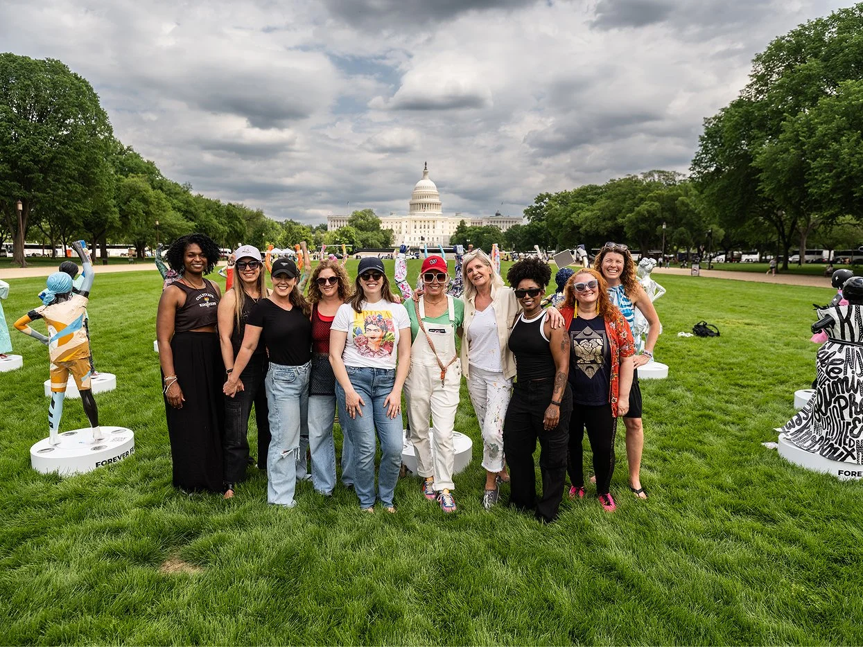 A group of ten women standing together on a grassy lawn in front of the U.S. Capitol building, with artistic sculptures around them and cloudy sky overhead.