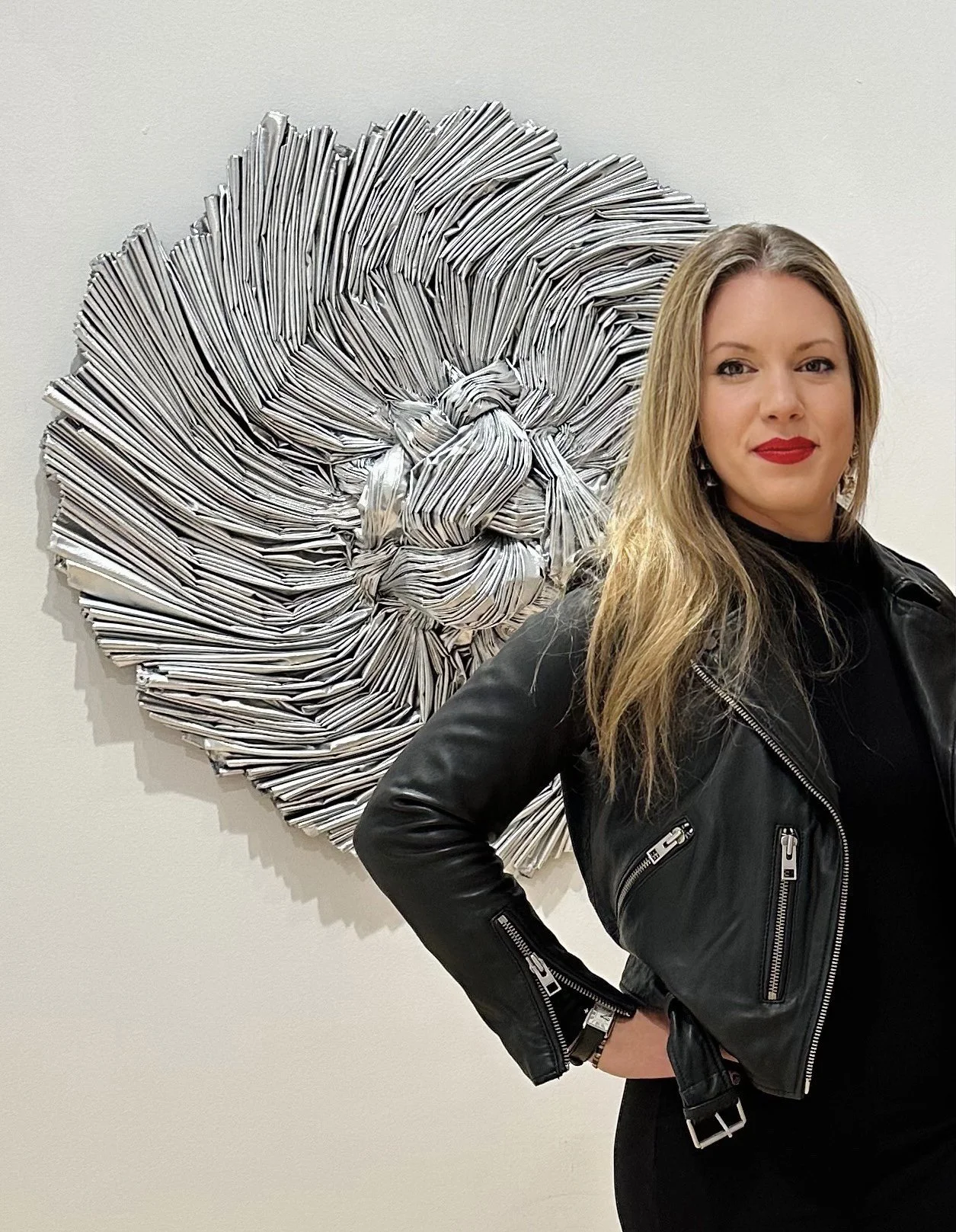 Woman with long blond hair wearing a black leather jacket standing in front of a large metallic art piece resembling a lion's mane.