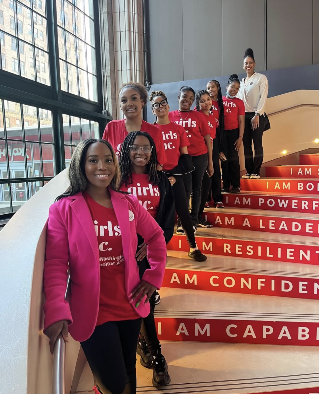 A group of women standing on a staircase with motivational phrases, with some wearing red shirts with 'girls dc' logo and one woman in a pink jacket, inside a building with large windows and city view.