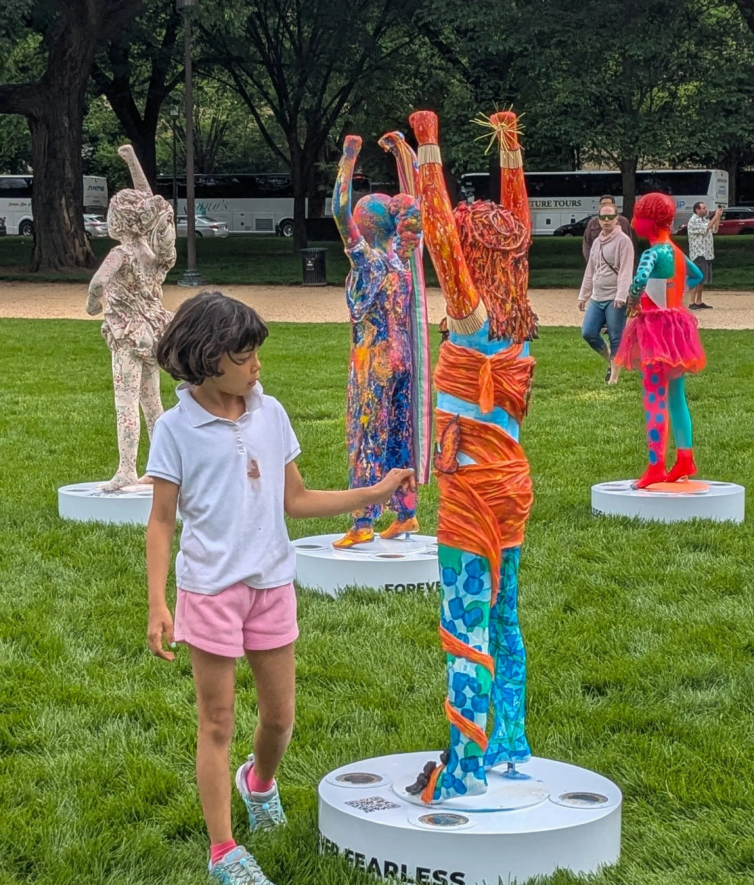 Child holding a colorful, abstract-patterned sculpture in an outdoor park with trees, people, and tour buses in the background.