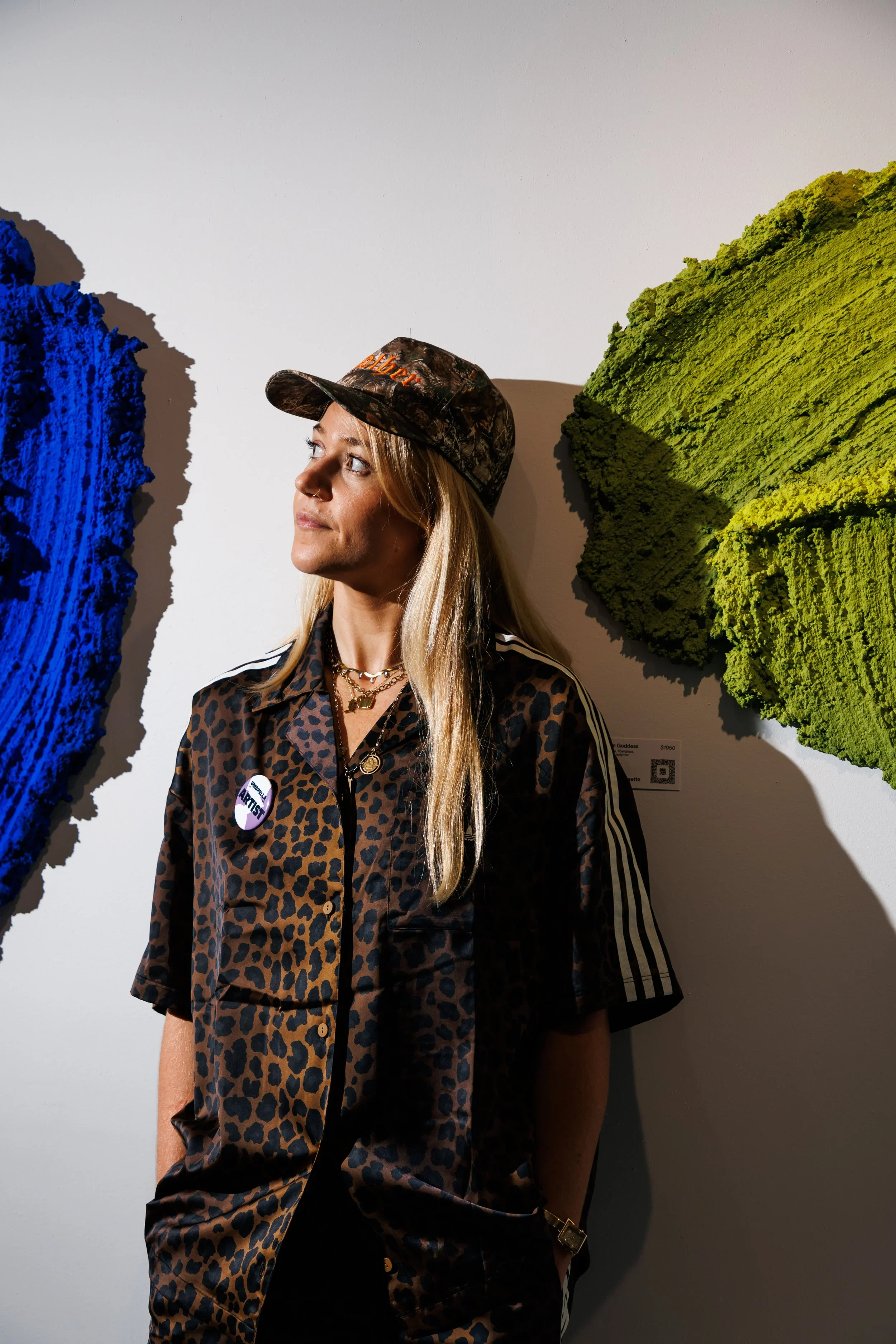 A woman wearing a leopard print jacket, gold jewelry, and a camo hat with orange text, standing in front of colorful textured art pieces on a plain white wall.