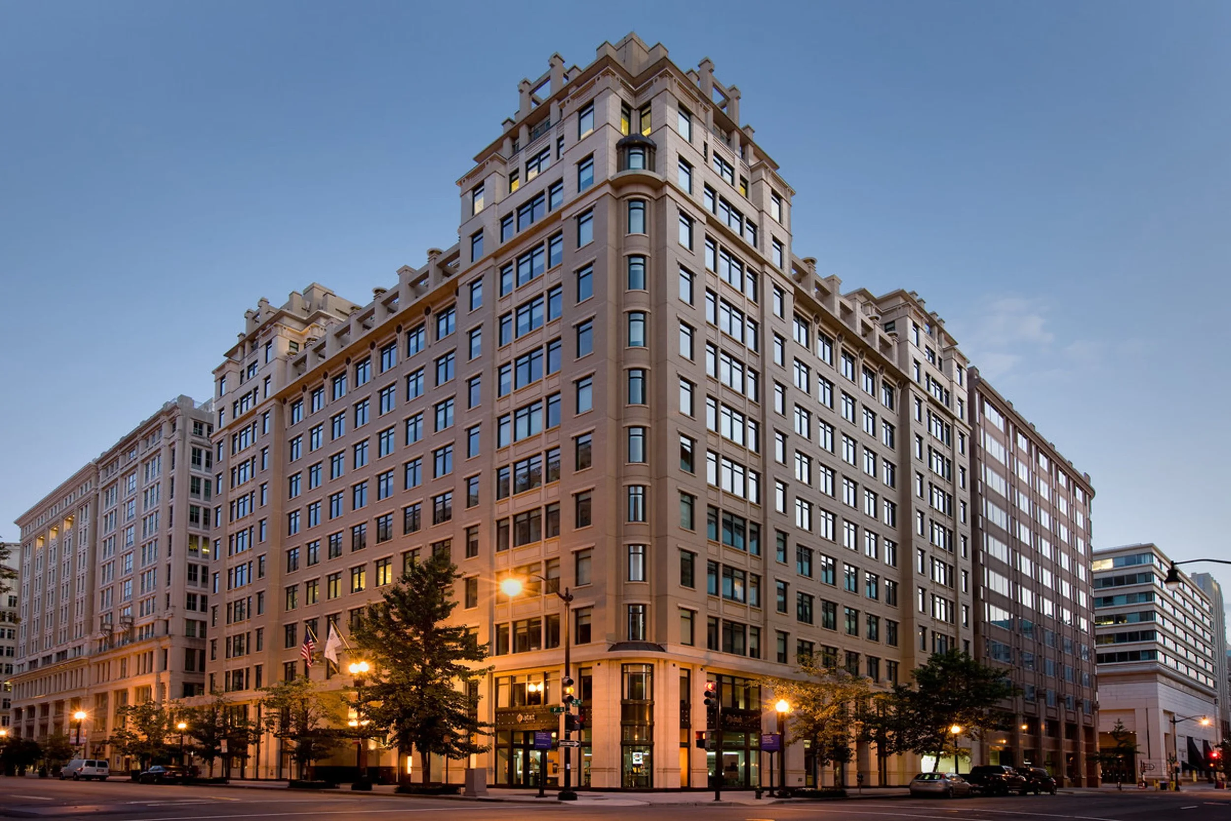 A tall beige-colored building with many windows, situated at a street corner, with trees and streetlights in the foreground and a clear blue sky overhead.