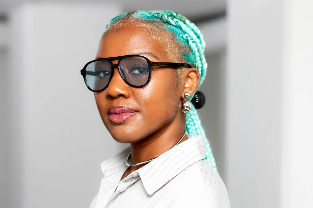 A woman with turquoise braided hair, wearing black sunglasses and pearl earrings.