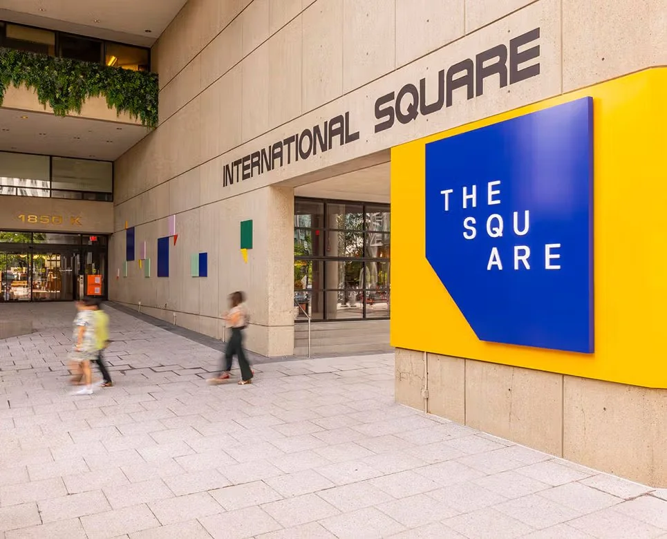 Entrance to International Square building with large sign reading 'The Square' in blue and yellow, with three children walking nearby.