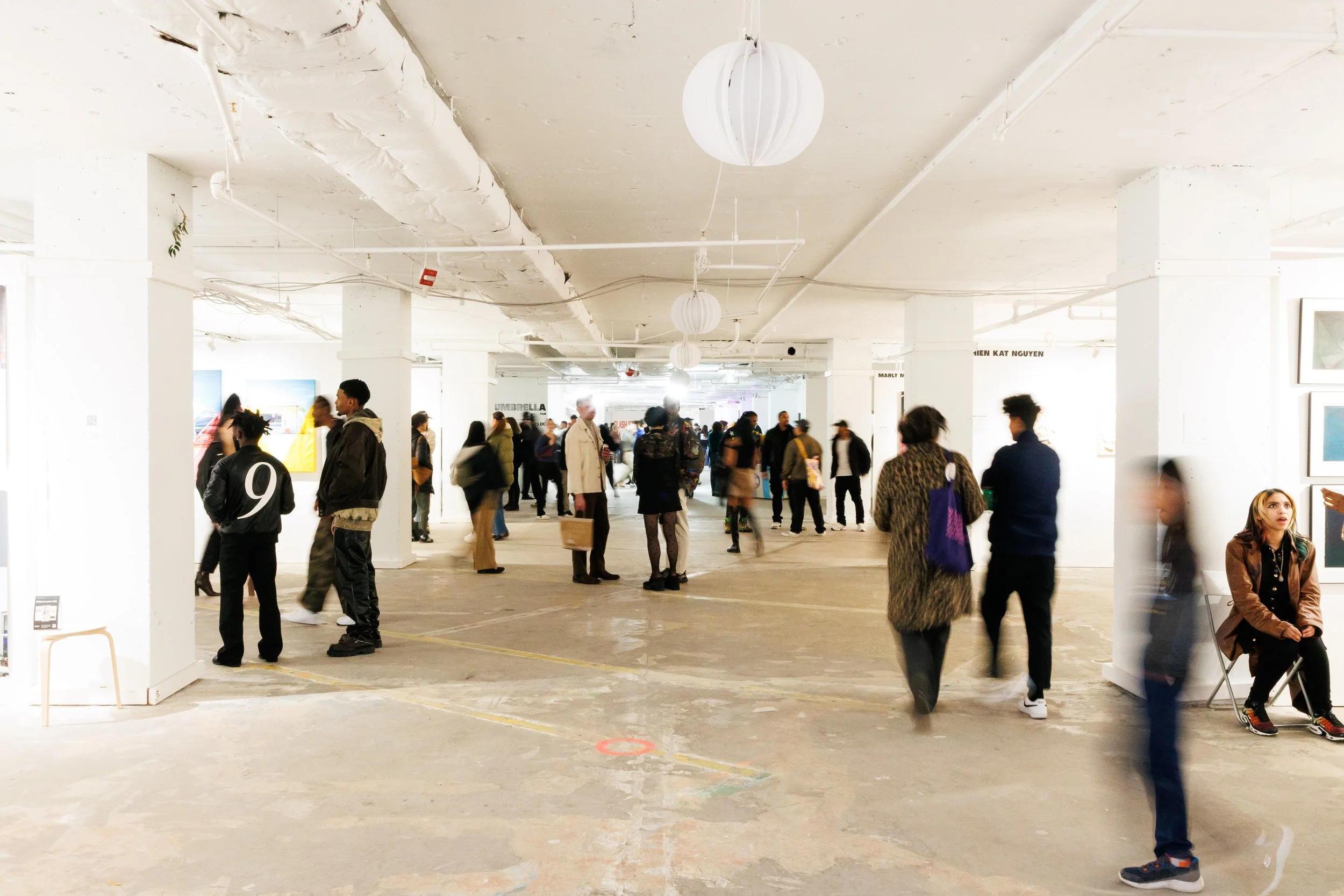 A busy indoor art gallery or exhibition space with many people walking, standing, and sitting, some looking at artworks. The room has white walls, exposed pipes, and decorative paper lantern ceiling lights.