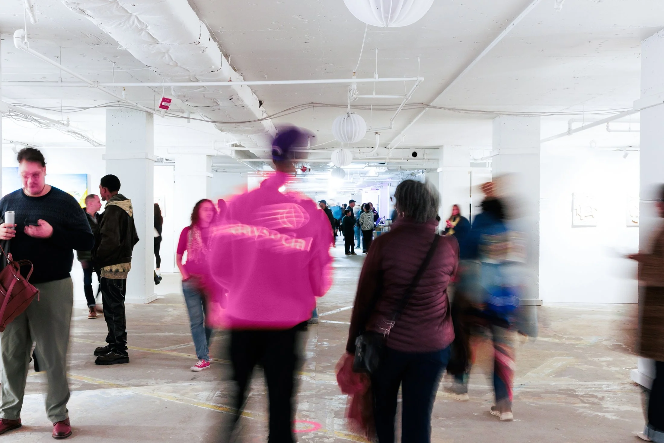 A crowded indoor space with people walking and talking. The ceiling is white with exposed pipes and hanging paper lanterns. The floor is concrete, and some people are blurry due to movement.