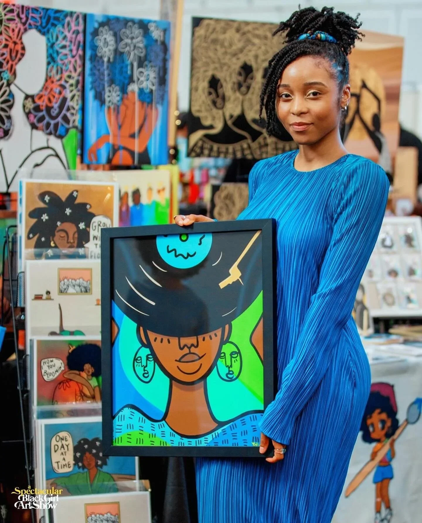 A young woman with braided hair in a bun wearing a blue pleated dress is standing in front of colorful artwork, holding a framed painting of a woman with earrings and a headscarf in a vibrant, stylized art style.