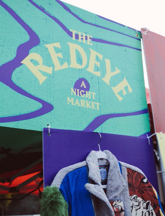 Sign for The Redeye, a night market, with a colorful background and clothing display underneath.