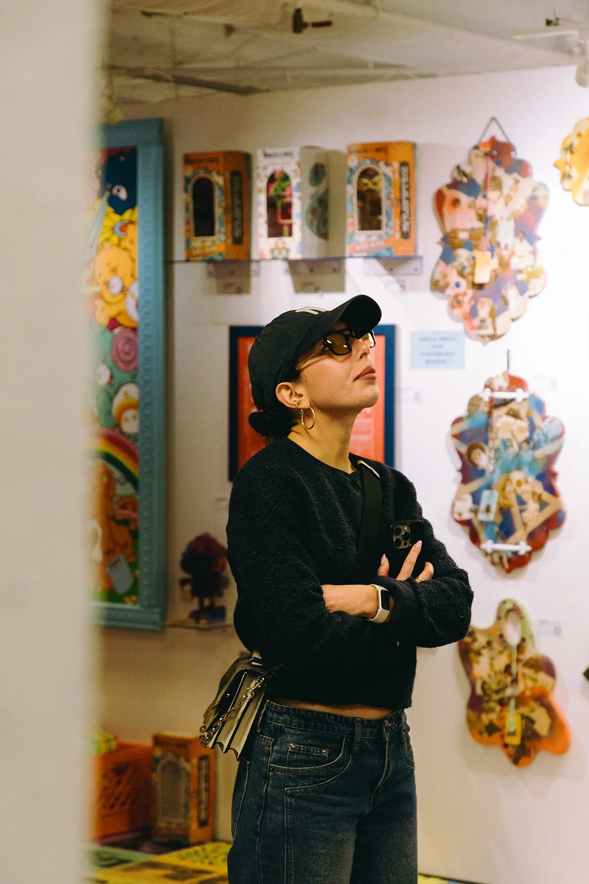 A woman with dark hair, wearing sunglasses, a black cap, and a black sweater, stands with arms crossed in an art gallery, looking up at colorful wall art.