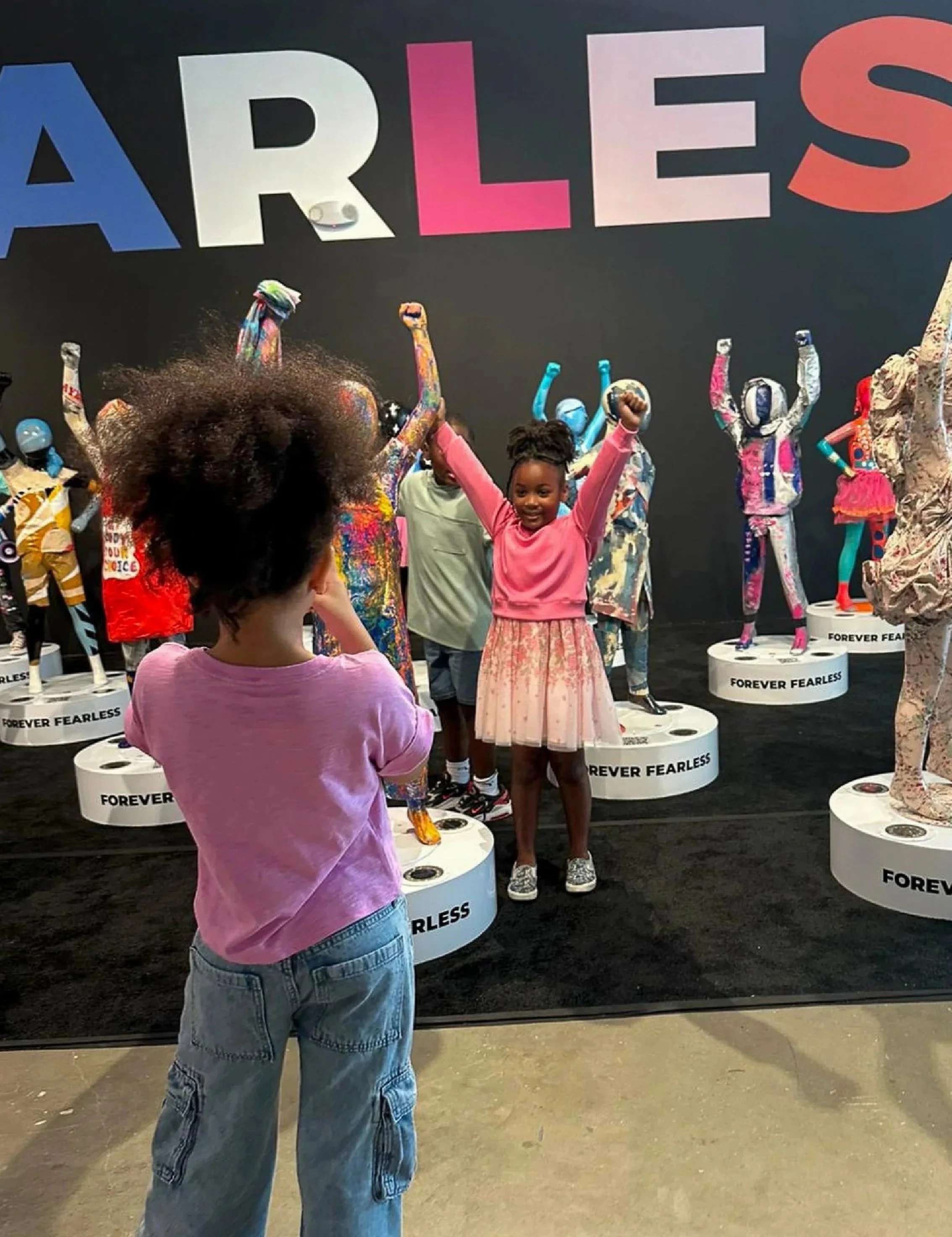 Children exploring mannequins dressed in colorful outfits at an exhibit with a large sign reading 'ARTLESS' and 'FOREVER FEARLESS' displayed on round platforms.