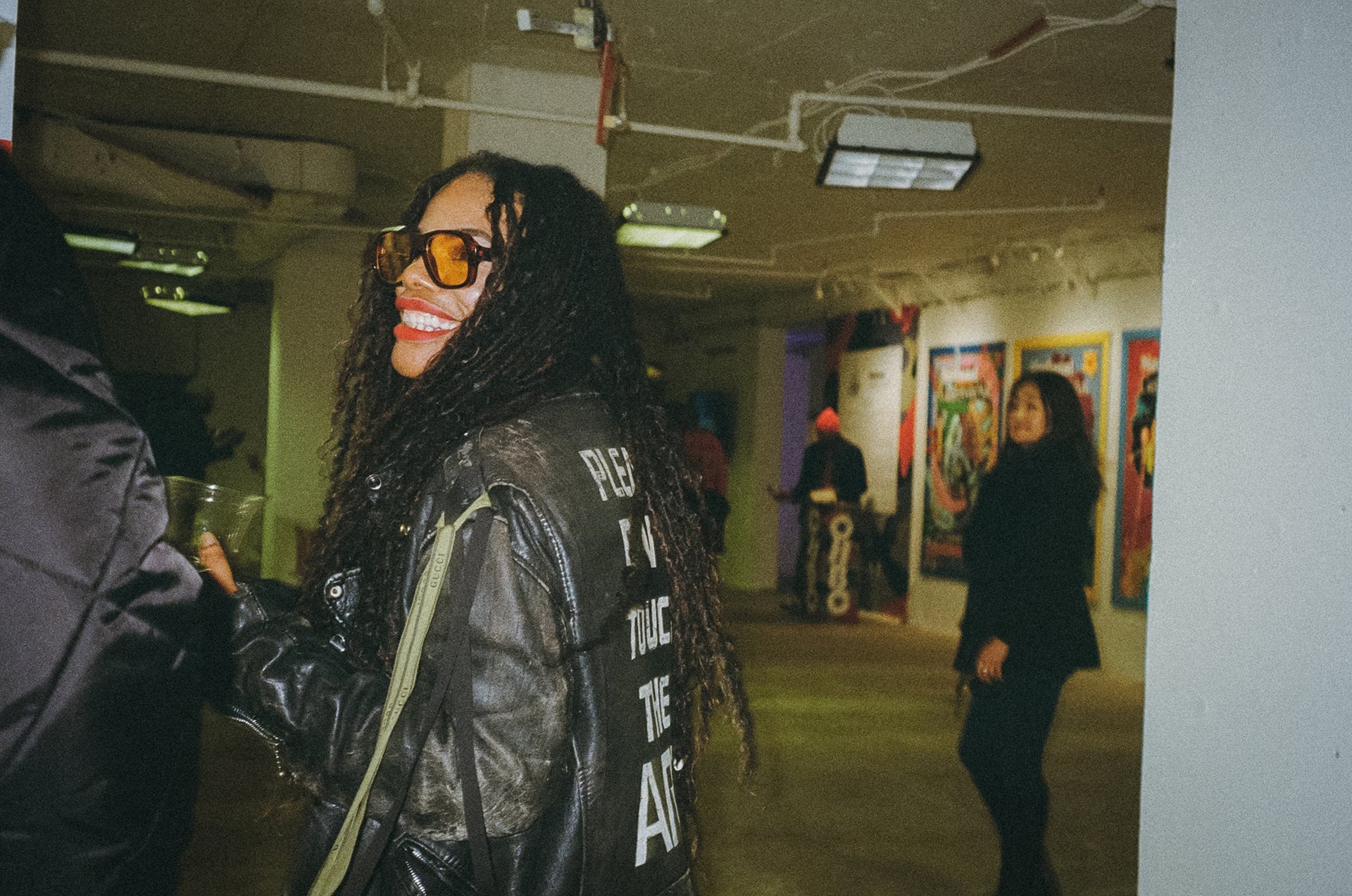 Smiling woman with long dreadlocks wearing sunglasses and a black jacket, standing in an art gallery, with another person partially visible on the left and a woman in black walking in the background.
