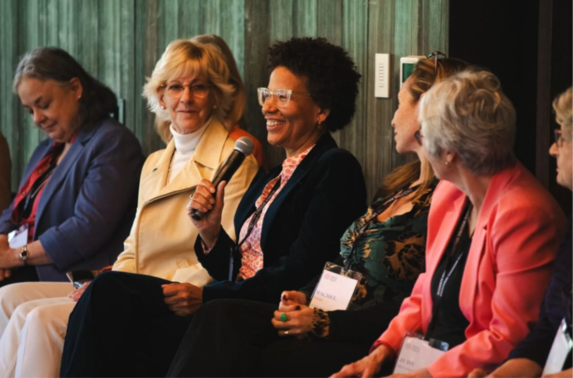 Women sitting on a panel at a conference, with the woman in the center speaking into a microphone, dressed in a dark jacket and glasses, with others listening.