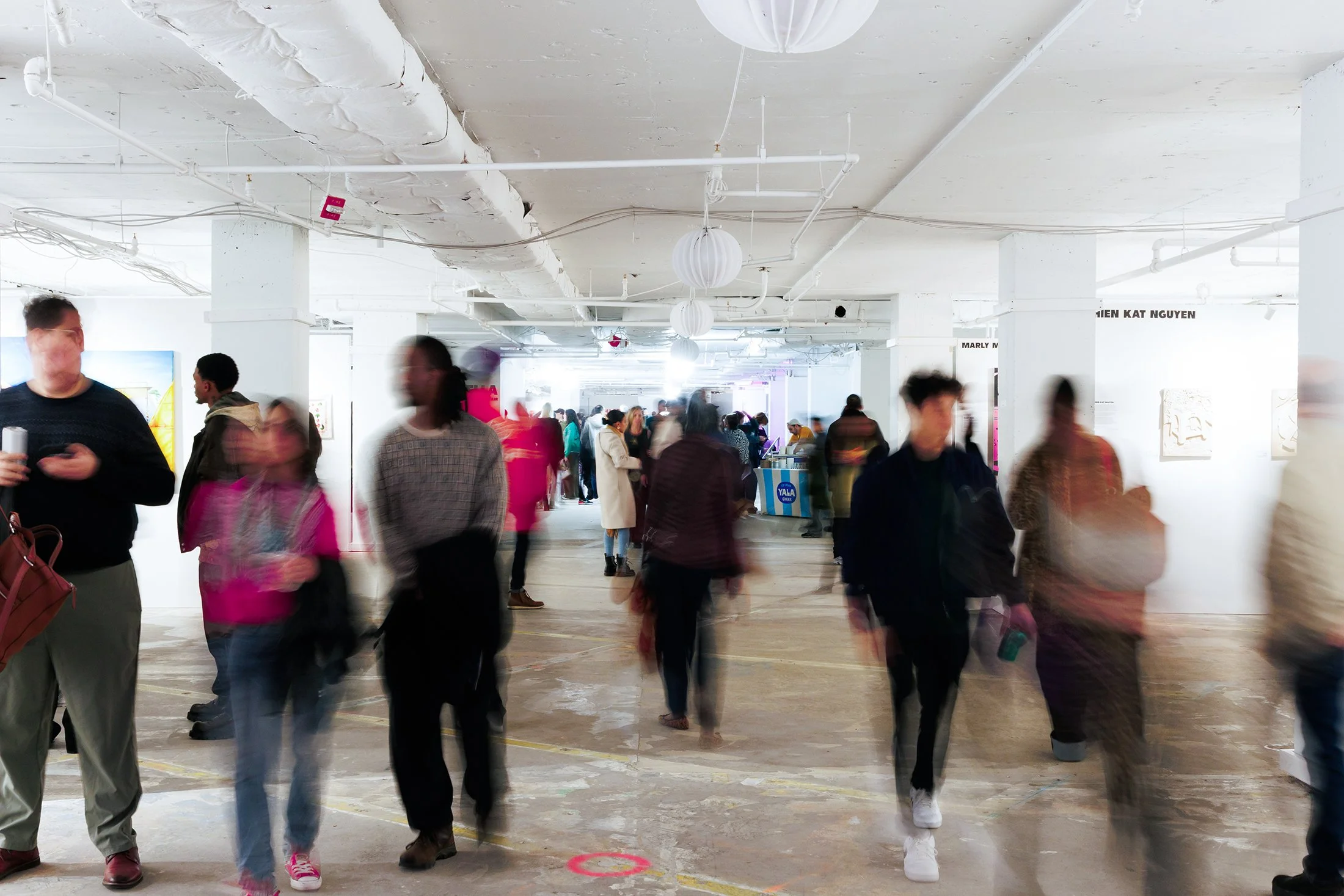 People walking through an art gallery with white walls and ceiling, featuring hanging paper lanterns and artwork. Some are blurred due to motion, indicating movement.
