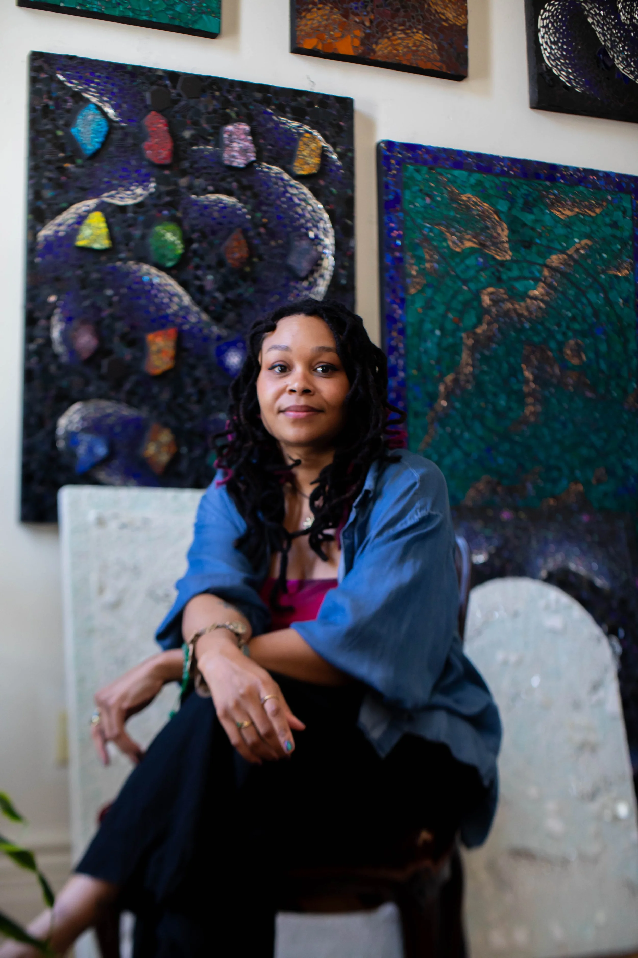 A woman with long dark curly hair sits in front of abstract paintings on a gallery wall. She is wearing a dark blazer over a pink top and has her arms crossed on her lap, looking at the camera.