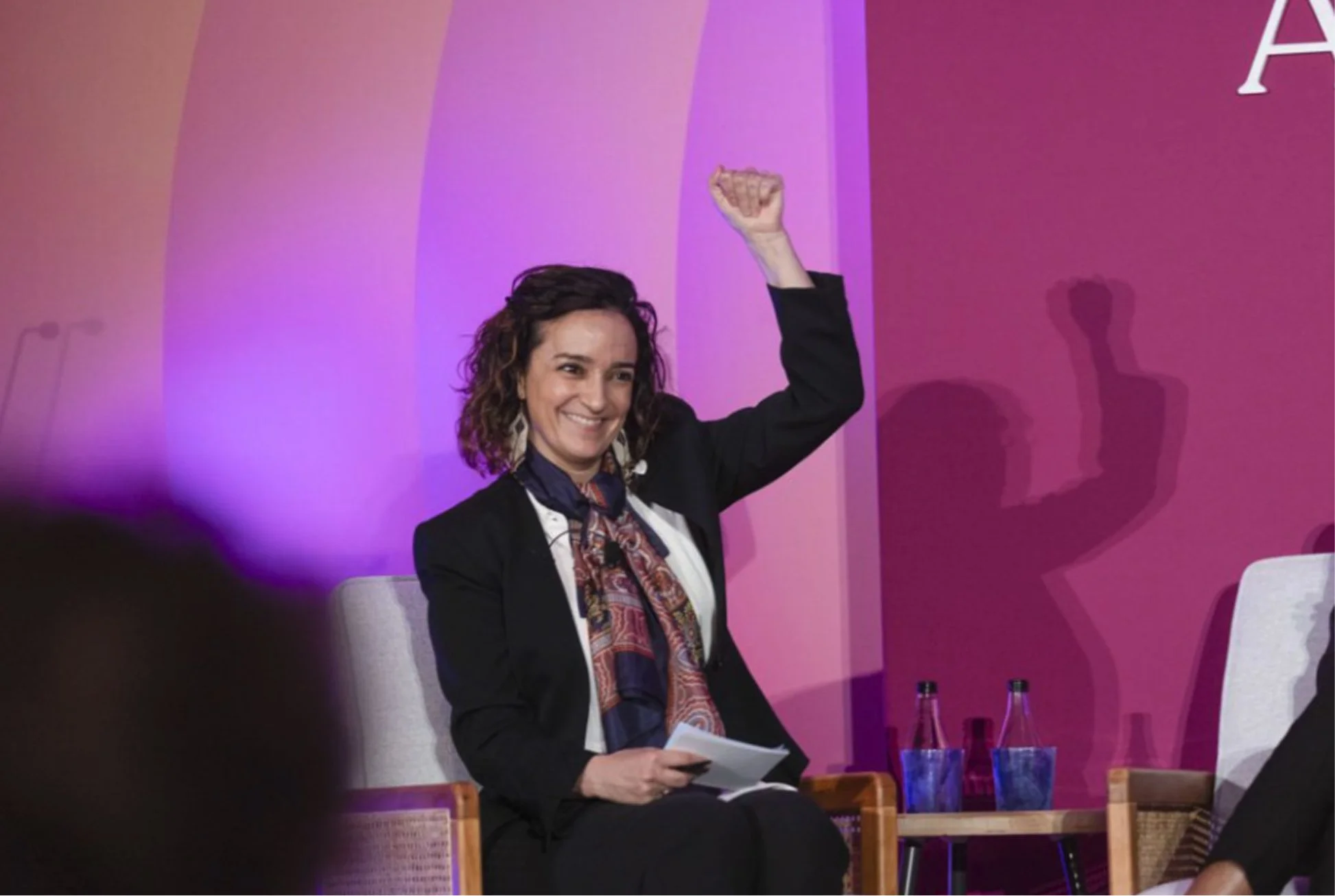 Woman with curly hair wearing a dark blazer and a patterned scarf, sitting on a chair on a stage with purple and pink background, raising her right fist in the air and holding papers in her left hand. There are three water bottles on a small table nearby.