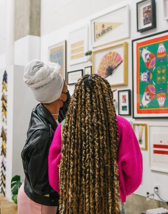 Two women viewing artwork in an art gallery, one with a towel wrapped around her head and wearing a black leather jacket, the other with long blonde dreadlocks and a bright pink sweater.