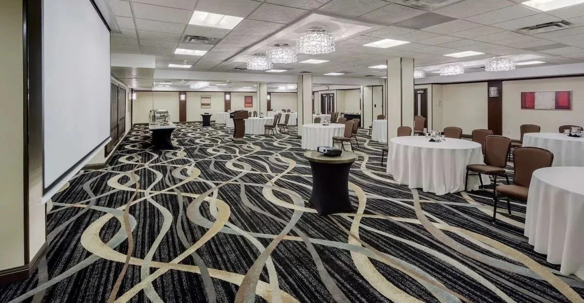 Empty conference or banquet room with round tables covered in white tablecloths, brown chairs, and a patterned carpet. There is a large screen on one side and chandeliers on the ceiling.