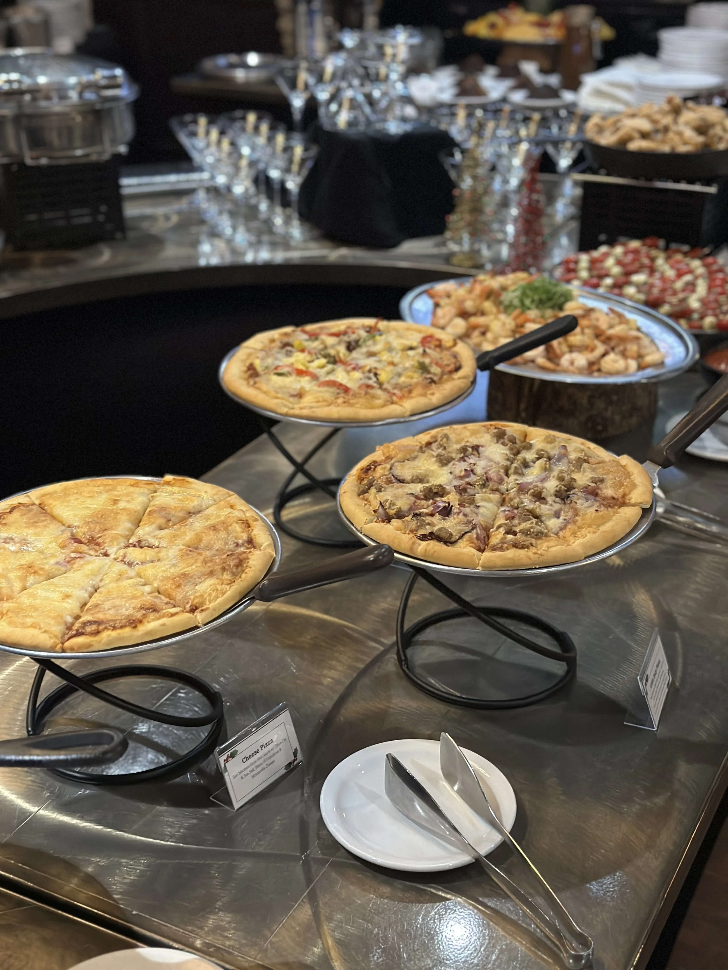 Assorted pizzas on display at a buffet table with other dishes and utensils in the background.