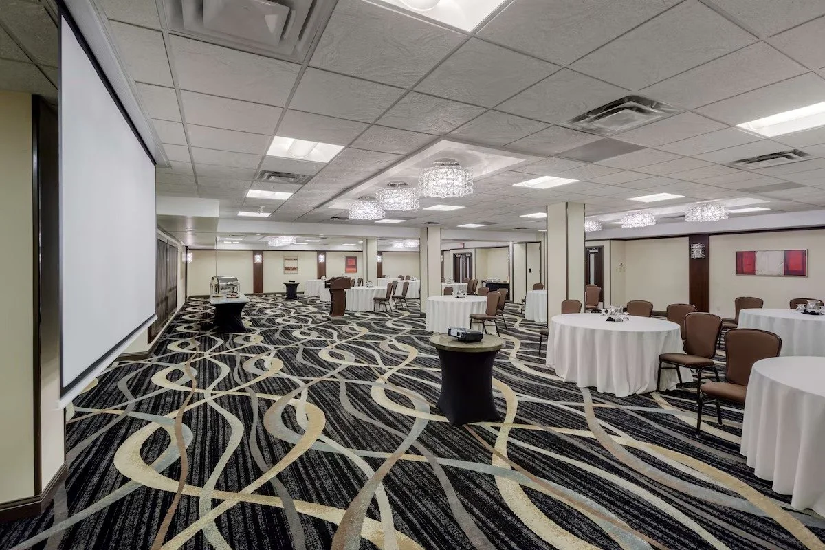 An empty banquet hall with round tables covered in white tablecloths, brown chairs, a large projection screen, and a patterned carpet. There are chandeliers and some artwork on the walls.