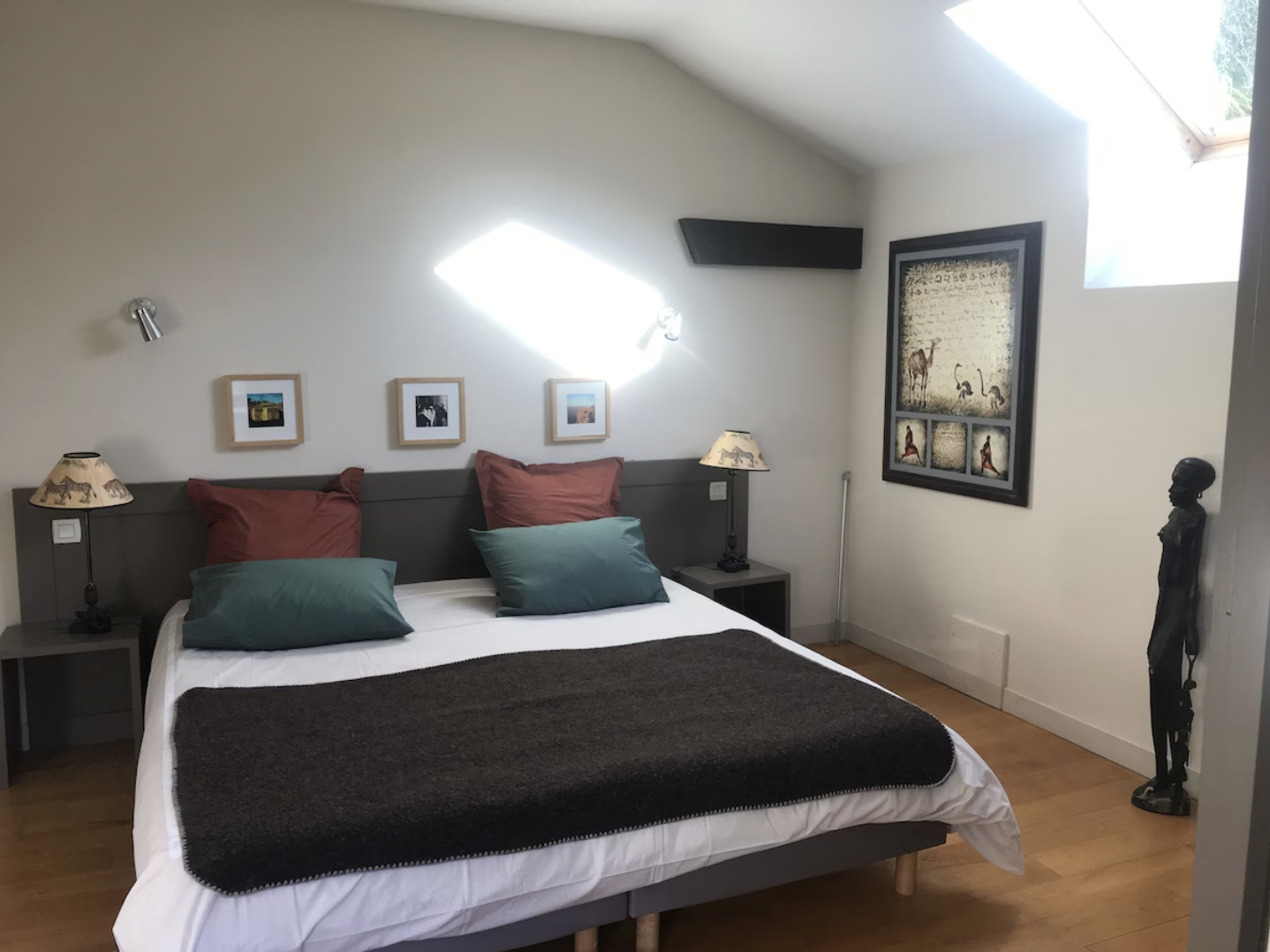 Chambre avec lit double, coussins vert et rouge, deux lampes de chevet, œuvres d'art sur les murs, sculpture en bois debout dans le coin droit.