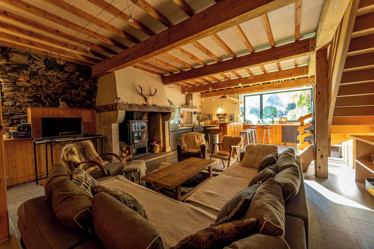 Salon de style rustique avec cheminée en pierre, canapés et fauteuils en tissu, table en bois, espace ouvert avec cuisine en arrière-plan, grande fenêtre laissant entrer de la lumière naturelle, plafond en bois avec poutres apparentes, décoration chaleureuse et cosy.