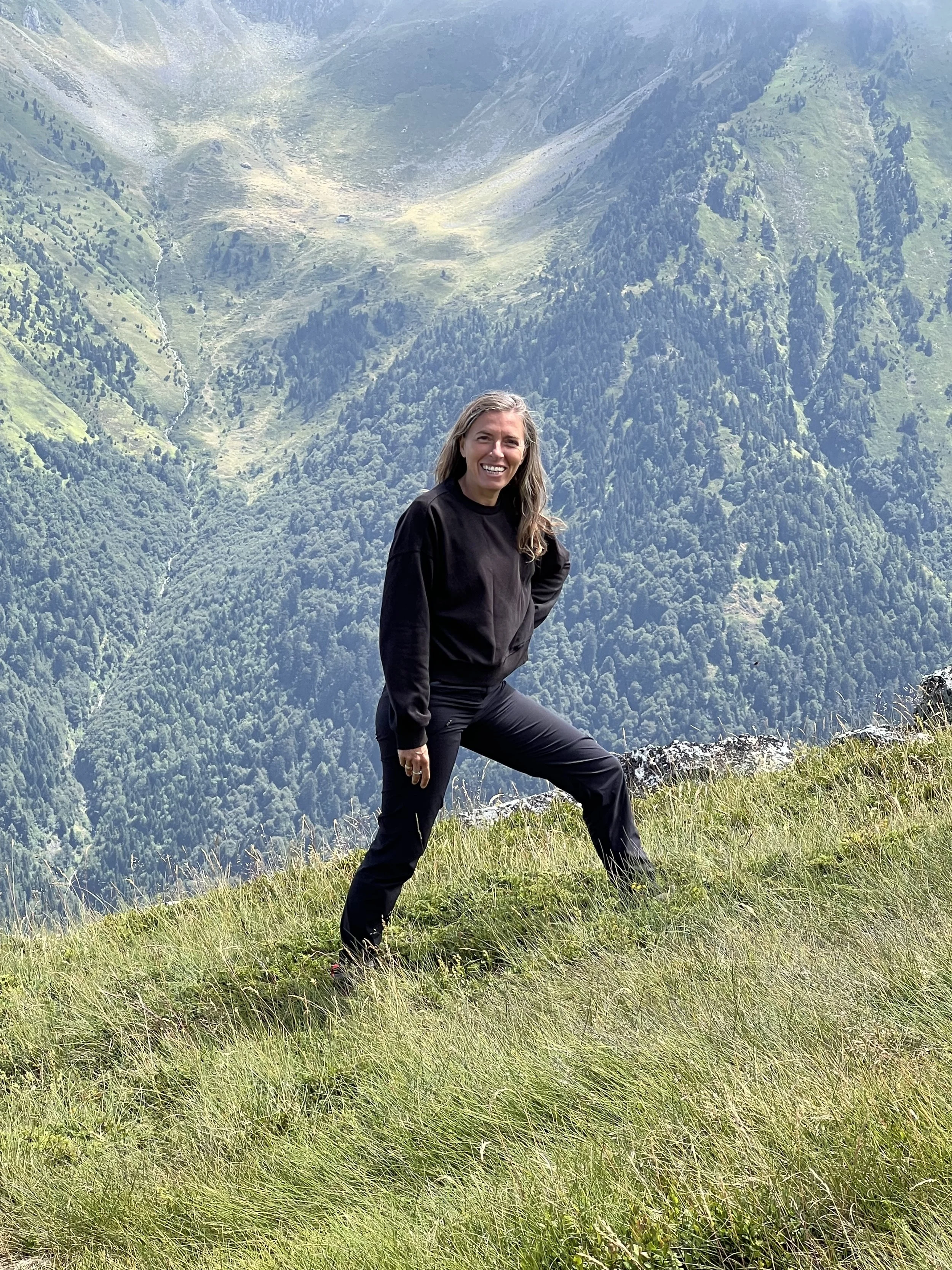 Une femme souriante en vêtements noirs, posant sur une colline enneigée avec un paysage montagneux en arrière-plan.