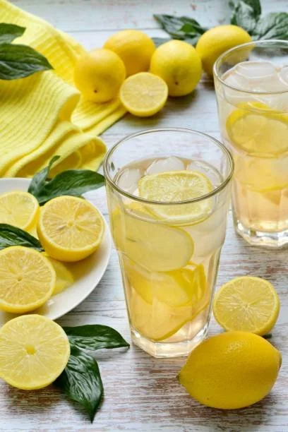 Verre de limonade avec des tranches de citron, citrons entiers et coupés, feuilles de menthe, et un linge jaune clair sur une table en bois.