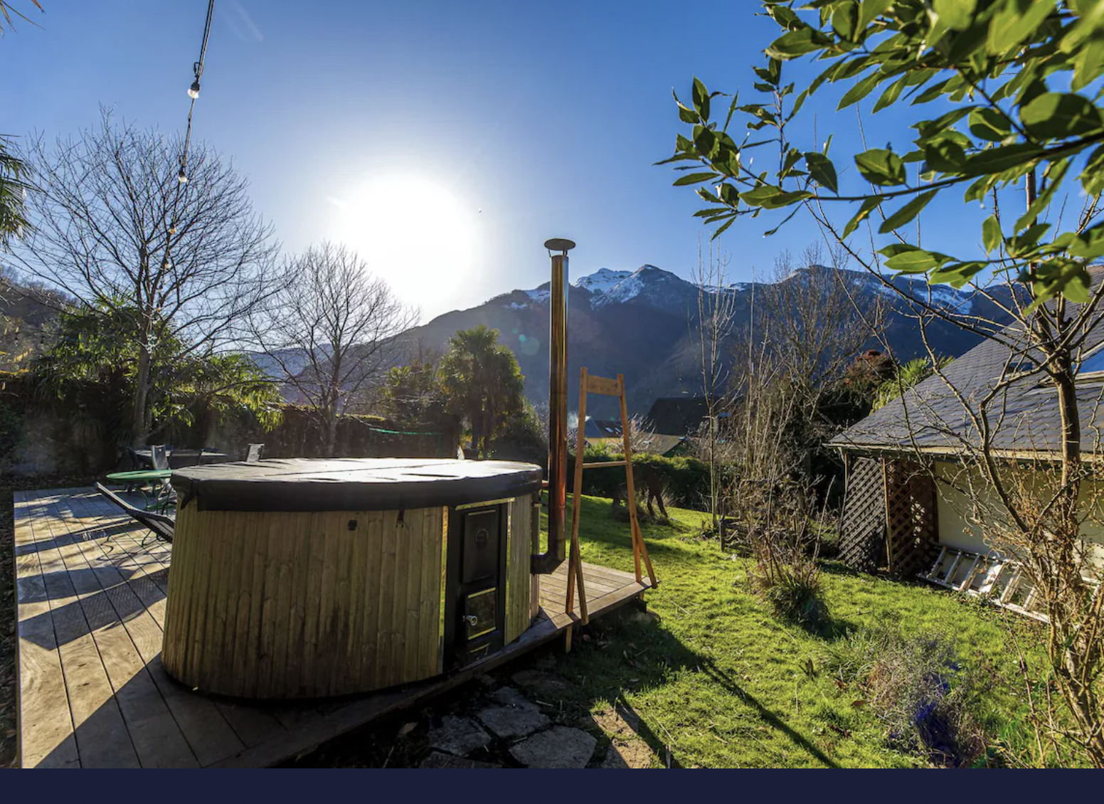 Jardin avec jacuzzi en bois, arbres sans feuilles, montagne enneigée en arrière-plan, ciel bleu ensoleillé.
