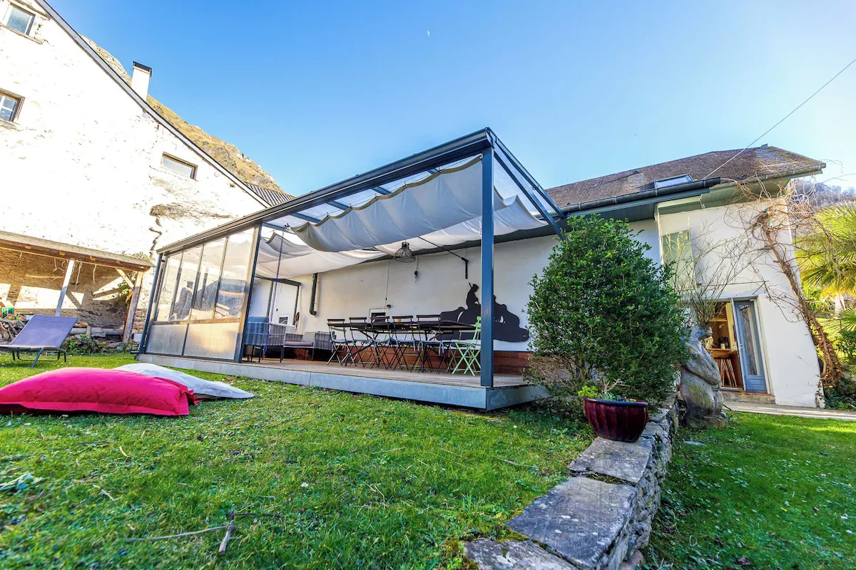Une terrasse en verre avec des meubles, un parasol et des coussins de sol dans un jardin ensoleillé, adjacent à une maison blanche avec un toit en pente.