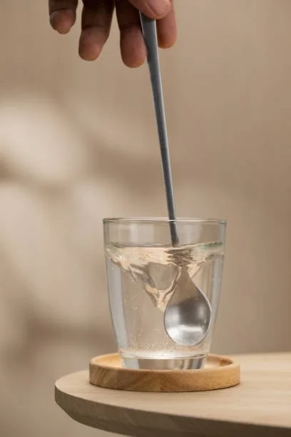Une main utilisant une cuillère pour mesurer un liquide dans un verre d'eau placé sur une petite assiette en bois.