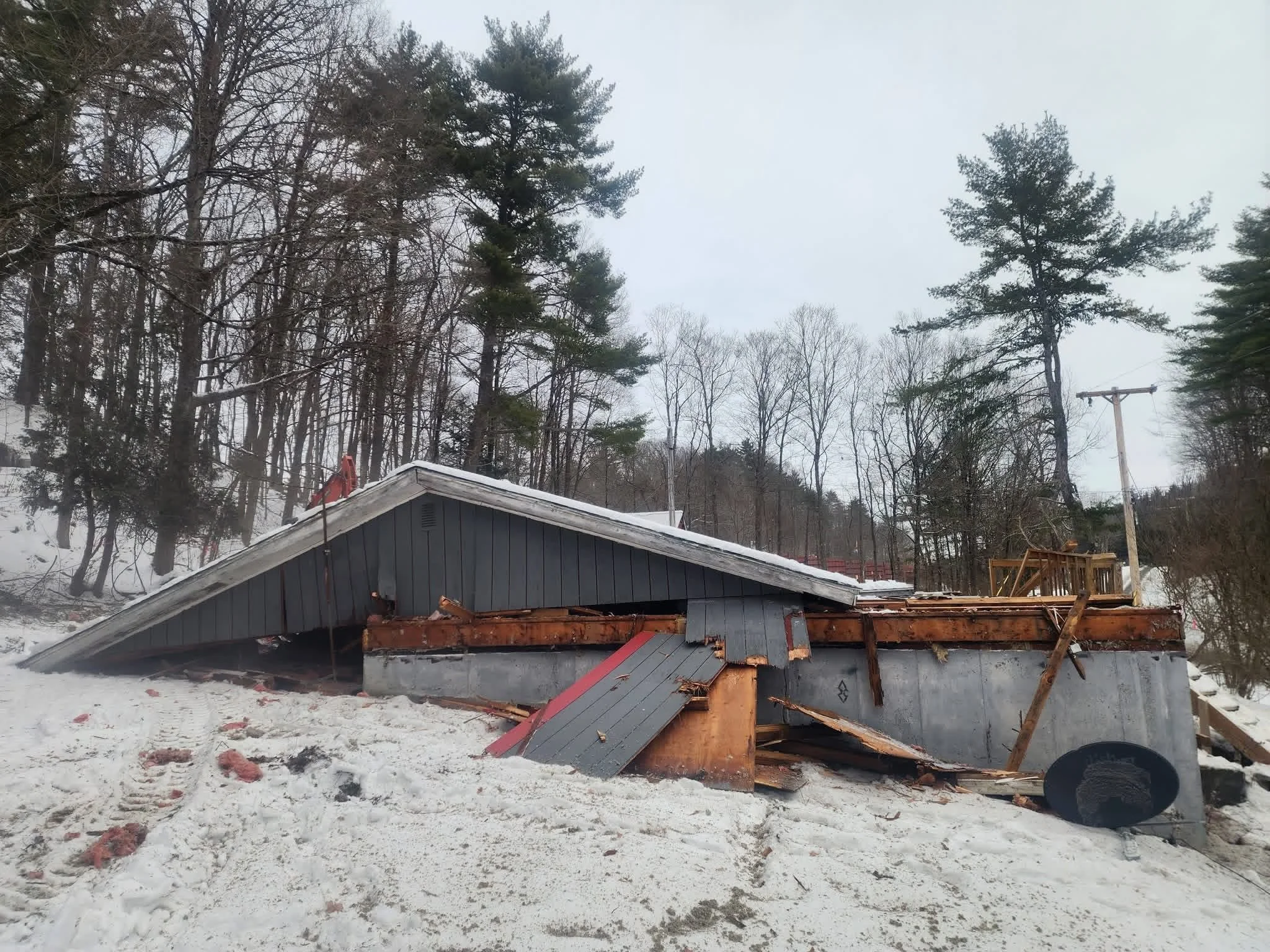 A house with a damaged roof and debris on the ground in a snowy yard, with trees and power lines in the background.