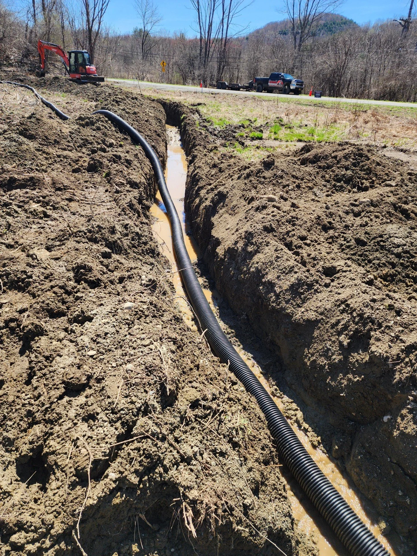A construction or utility repair site with a black corrugated pipe laid in a deep trench, with a backhoe excavator and a pickup truck parked nearby, with orange cones and warning signs on the side of the road amid a forested area with a mountain in the background.