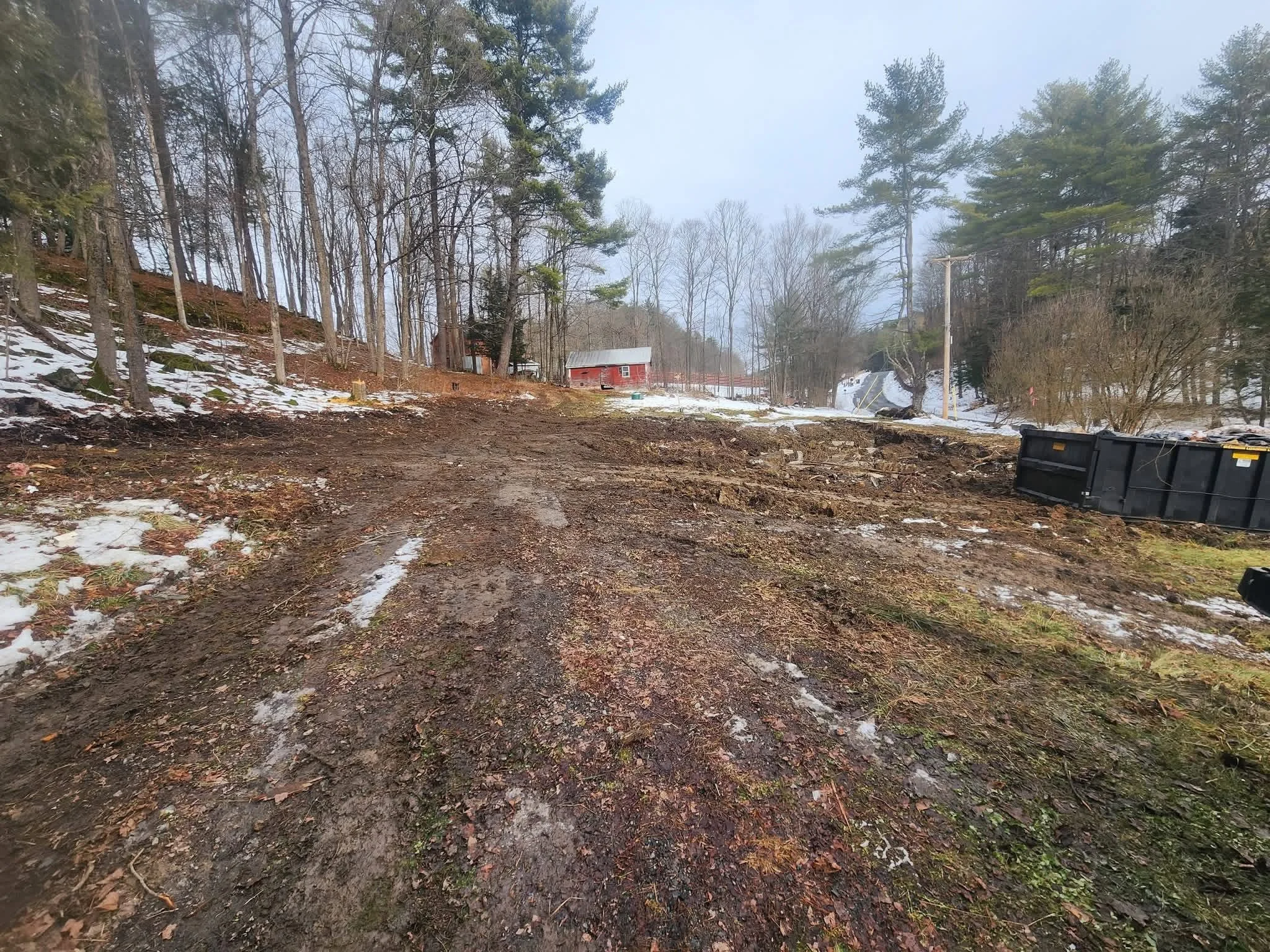 A muddy dirt road in a wooded area with snow patches, a red barn in the distance, and trees on both sides, with a cloudy sky overhead.