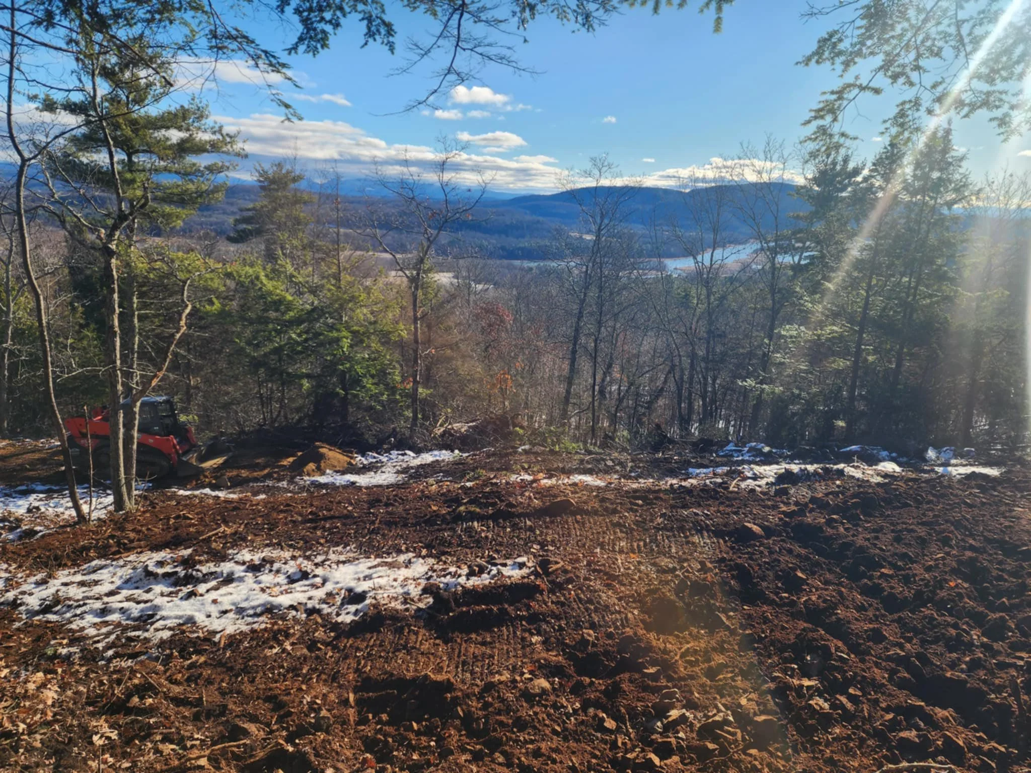 A scenic view of a mountainous landscape with a clear blue sky, scattered clouds, and trees in the foreground, some snow on the ground, and construction equipment partially visible.