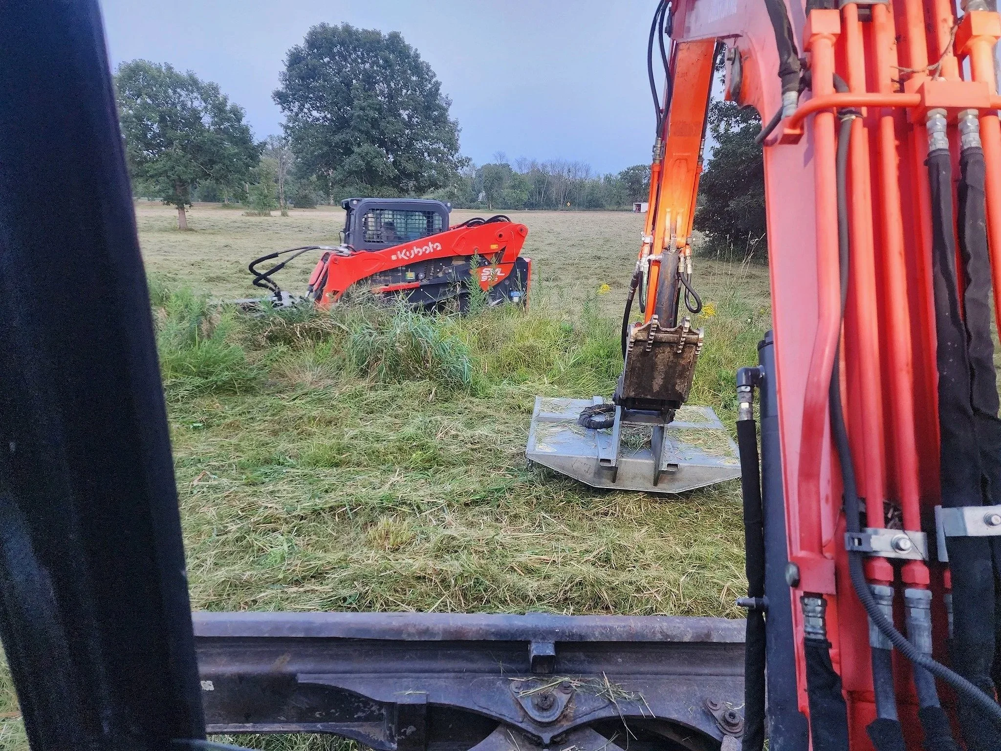 View from inside a construction vehicle showing an orange excavator and a grassy field with trees in the background.