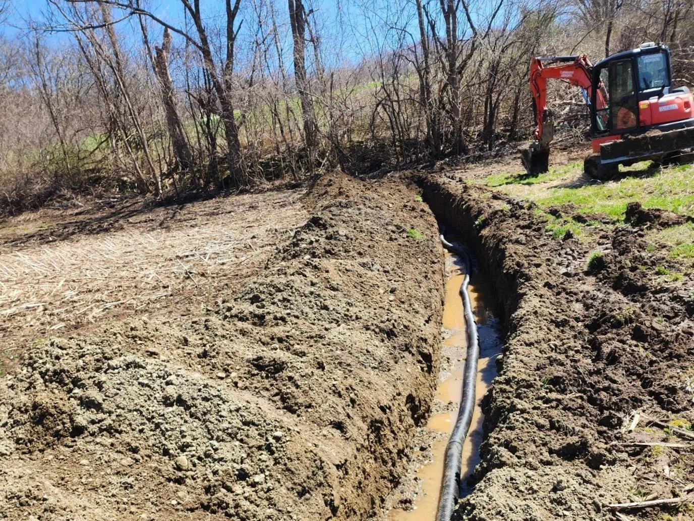 Excavation site with a narrow trench, a black pipeline installed inside, and a mini excavator on the right side amid trees and grass.