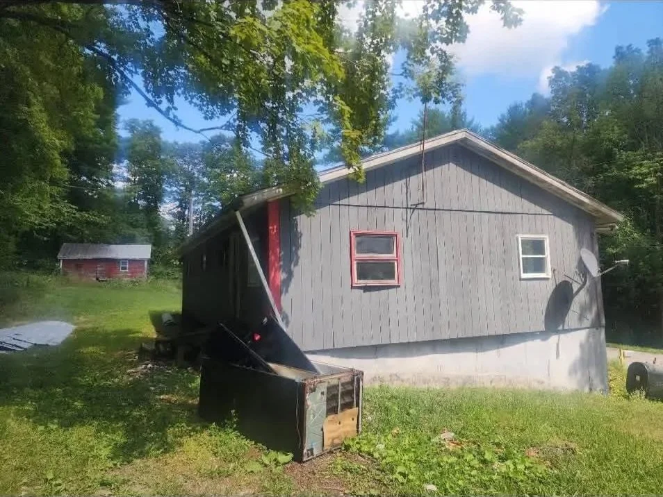 A gray house with red window frames and a satellite dish, surrounded by greenery and trees. There is a damaged black object in front of the house, and a smaller red shed in the background.